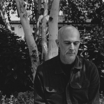 A black and white image of a white man in a dark casual shirt looking down. Behind him is a silver birch tree.
